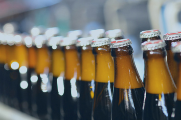 A row of brown glass beer bottles with metal caps moving along a production line in a brewery, with a blurred background.
