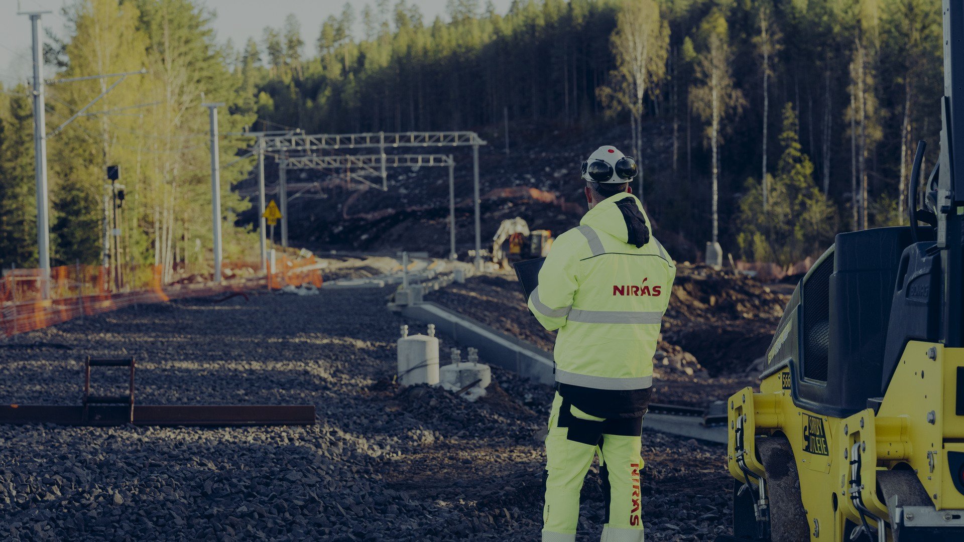 En ingeniør med reflekterende tøj observerer en jernbanebyggeplads omgivet af skov, med fokus på detaljer