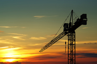 A large construction crane silhouetted against a colorful sunset sky with hues of orange, yellow, and blue.