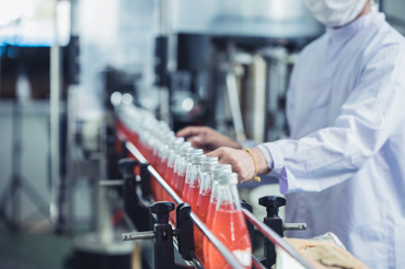 A factory worker wearing protective clothing and a mask is overseeing a production line where glass bottles filled with a red liquid are being processed. The setting suggests a controlled environment, possibly in a beverage manufacturing facility.