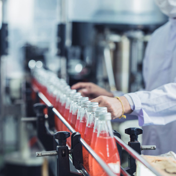 A factory worker wearing protective clothing and a mask is overseeing a production line where glass bottles filled with a red liquid are being processed. The setting suggests a controlled environment, possibly in a beverage manufacturing facility.